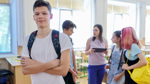 Im Vordergrund ist  ein junger Schüler zu sehen. Im Hintergrund sind zwei Schülerinnen, ein Schüler und die Lehrerin zu sehen, wie sie sich unterhalten. Copyright: Valerii Honcharuk | stock.adobe.com